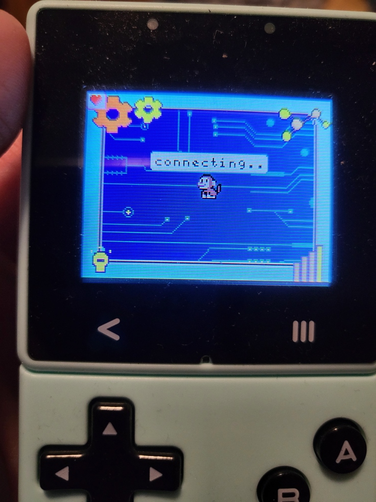 A photo of an Ovobot handheld game console, on the screen is pixel art of a blue "tech" background with colorful gears and circuit board wiring. In the middle is a small monkey that says "connecting.."